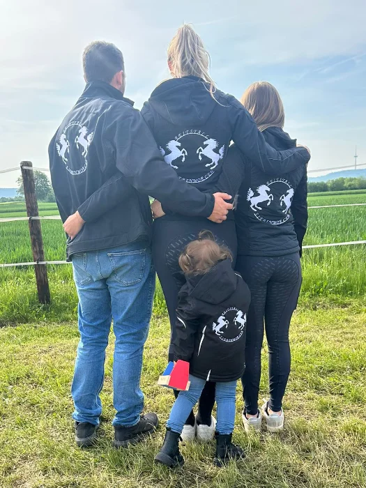 Personen mit schwarzen Jacken mit Reitstall Siebenmorgen Logo, die nebeneinander auf einer grünen Wiese stehen und in die Ferne blicken.