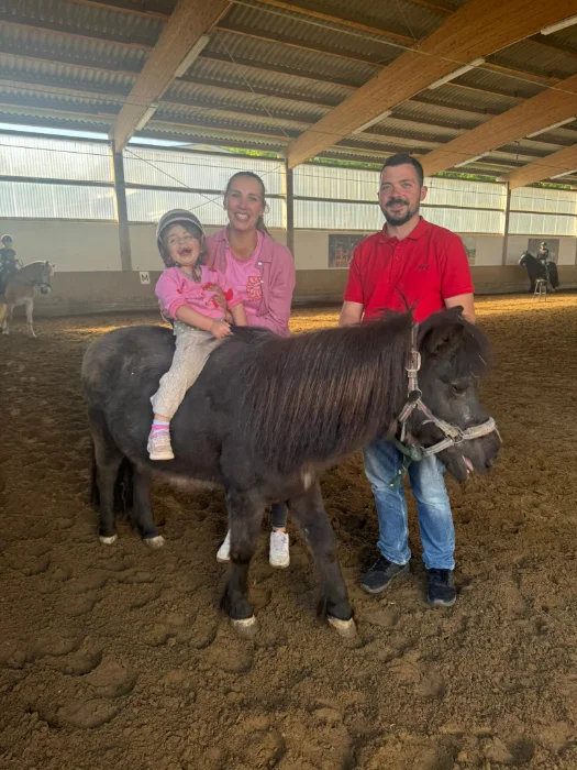 Ein Kind mit Helm sitzt lachend auf einem Pony, begleitet von zwei Erwachsenen in einer Reithalle.