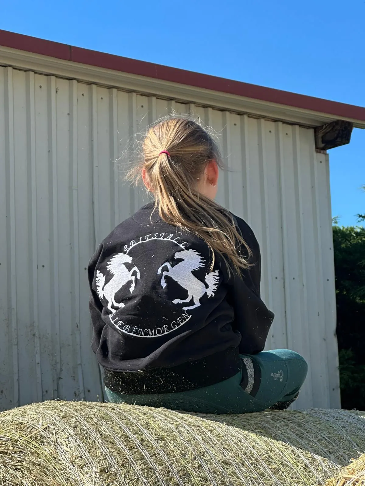 Mädchen mit Reitstall Siebenmorgen Jacke sitzt auf Heuballen vor einer Scheune bei sonnigem Himmel.